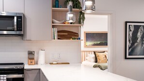 Ett modernt kök med en inbyggd mikrovågsugn, en spis och en vit bänkskiva i marmor. Det finns en hylla i trä med dekorativa föremål och en inramad bild av ett berglandskap.