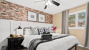 Ein Schlafzimmer mit Backsteinwand, einem Bett mit weißer und grauer Bettdecke, einem schwarzen Nachttisch mit Lampe und einem Fenster mit Vorhängen.