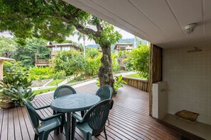 A wooden deck with a table and chairs, surrounded by lush greenery and a view of buildings in the background. - Bangalô 08 - Ilhabela (Ilhabela)
