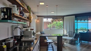 A modern kitchen with a central island, a dining area with a view, and a cozy seating space. - Bangalô 08 - Ilhabela (Ilhabela)