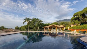 A swimming pool with lounge chairs and a gazebo, surrounded by greenery and mountains. - Bangalô 08 - Ilhabela (Ilhabela)