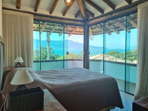 A bedroom with a large bed, a bedside table with a lamp, and a view of the sea and mountains through large windows. - Bangalô 08 - Ilhabela (Ilhabela)