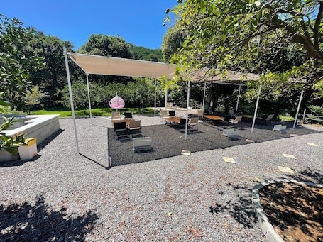 An outdoor seating area with tables and chairs under a pergola, surrounded by gravel and greenery.