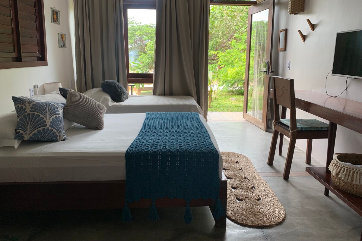A bedroom with a bed, a desk, and a TV. There is a window with curtains and a view of greenery outside.
