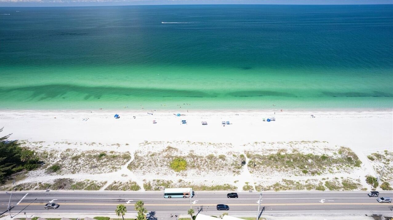 A beach with clear turquoise water, a sandy shore, and a road with vehicles.