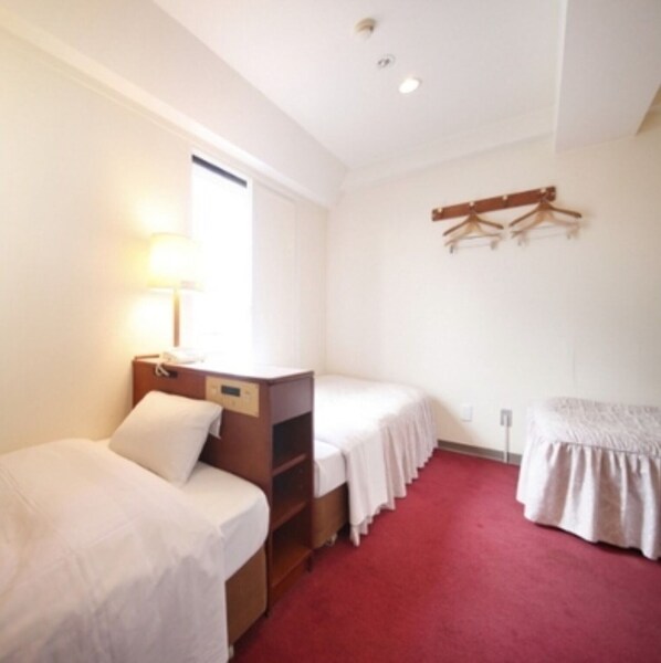 A hotel room with two single beds, a desk with a chair, a bedside table, a lamp, and a coat rack.