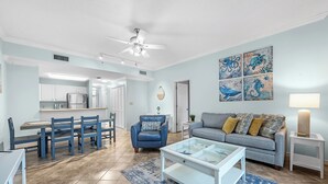 A modern living room with a blue sofa, a glass coffee table, and a dining area with blue chairs. - 401 Dunes at the end of Fort Morgan Road (Gulf Shores)
