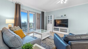 A modern living room with a glass coffee table, a sofa, a television, and a ceiling fan. - 401 Dunes at the end of Fort Morgan Road (Gulf Shores)