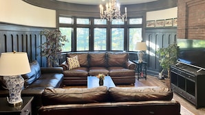 A living room with a brown leather sofa, a coffee table, a chandelier, and a television. - Spectacular Historic Mansion w/Pool&Tennis (Goshen)