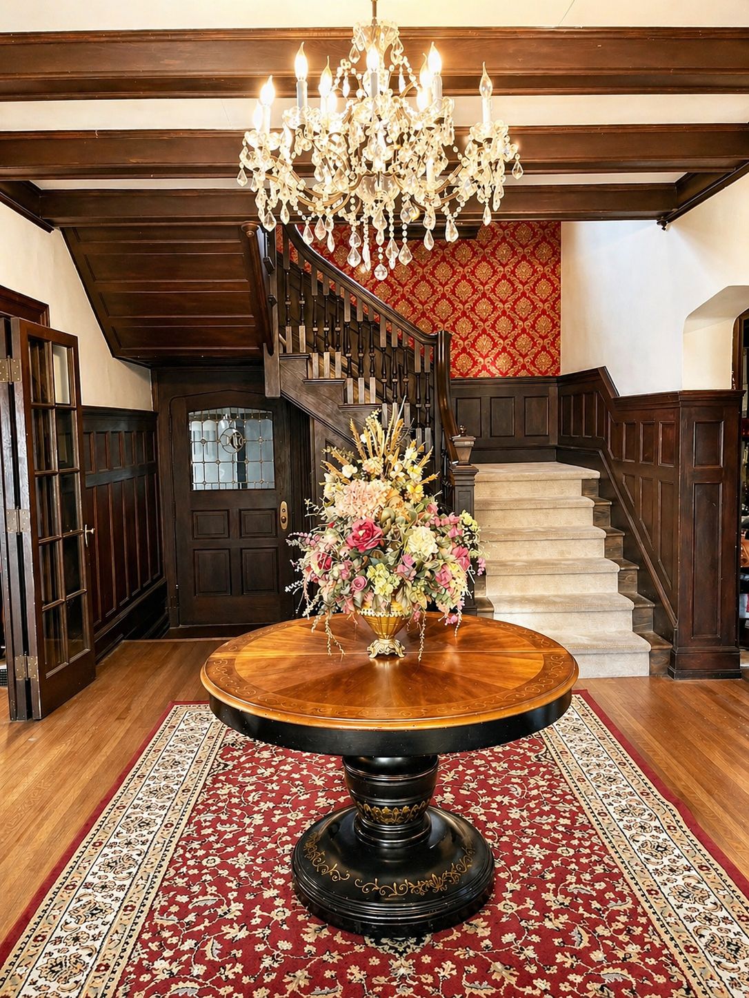 A grand staircase with a chandelier, a round wooden table with a floral centerpiece, and a patterned carpet.