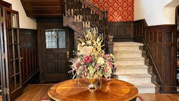 A grand staircase with a chandelier, a round wooden table with a floral centerpiece, and a patterned carpet.