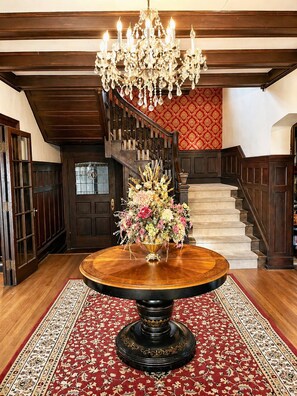 A grand staircase with a chandelier, a round wooden table with a floral centerpiece, and a patterned carpet. - Spectacular Historic Mansion w/Pool&Tennis (Goshen)