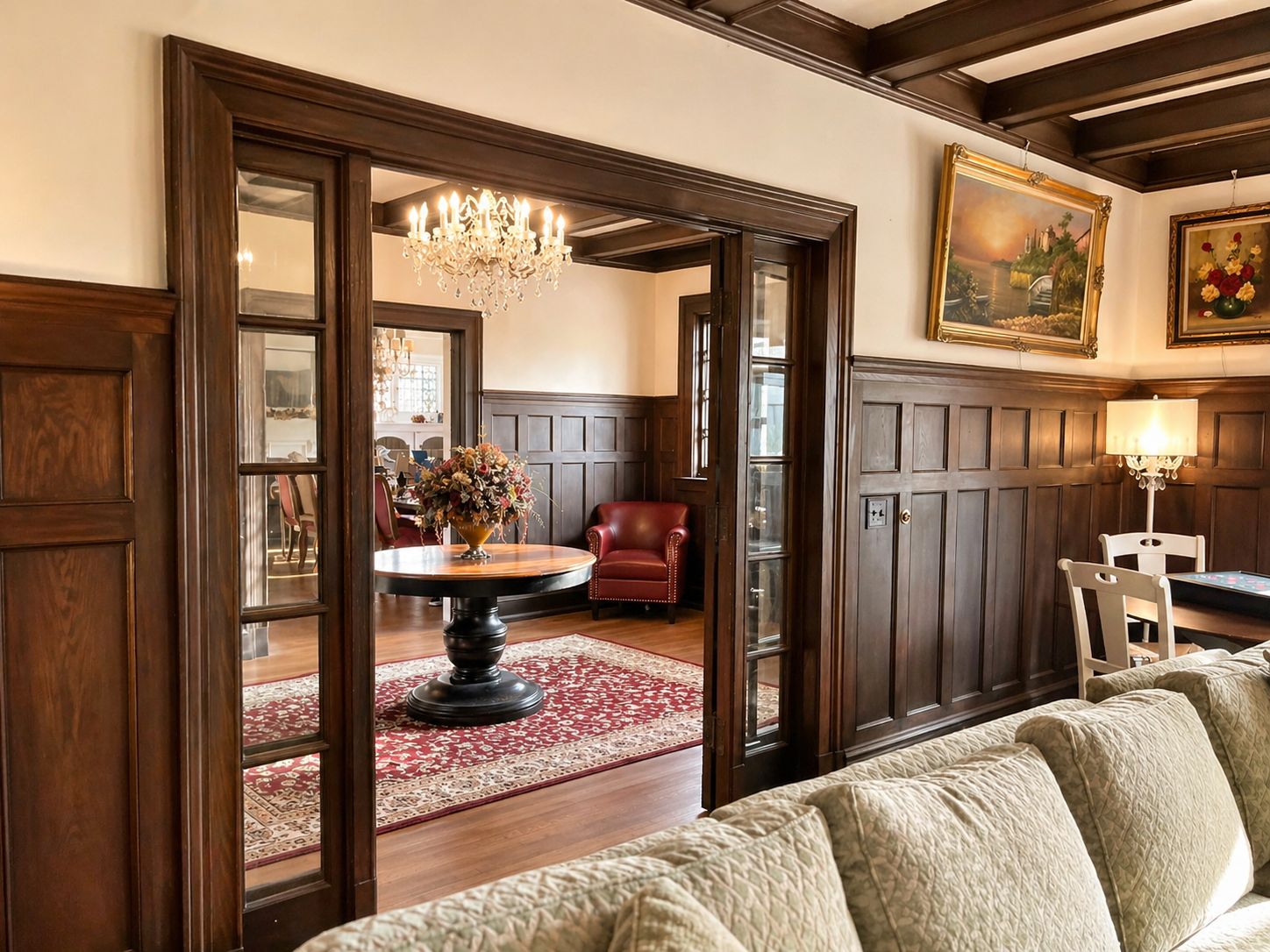 A room with a round table, a red leather chair, a sofa, a chandelier, and two paintings on the wall.