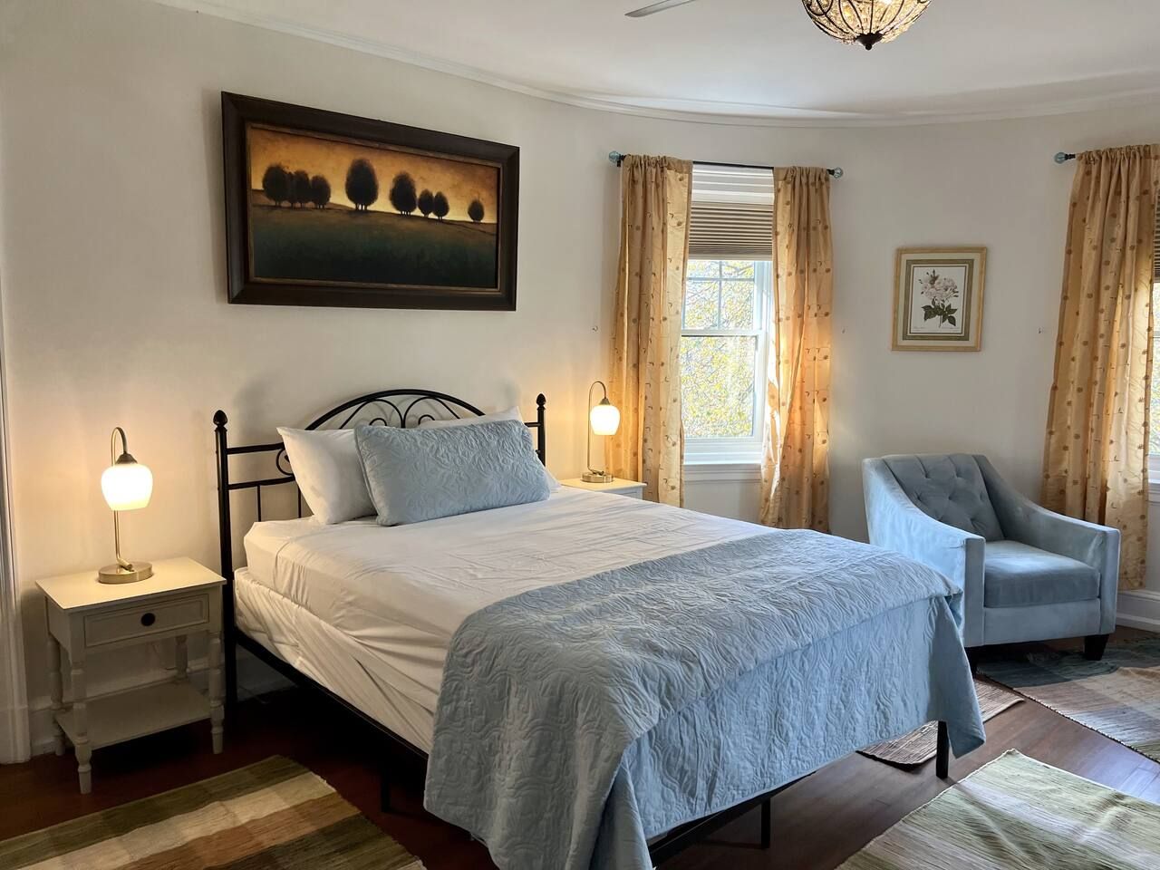 A bedroom with a bed, bedside tables, a chair, a painting, and a window with curtains.