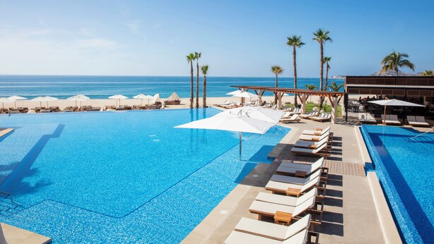 A pool area with lounge chairs and umbrellas, palm trees, and a view of the ocean. - Altitude by Krystal Grand Los Cabos – All Inclusive (San José del Cabo)