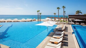 Zona de piscina con sillones reclinables y sombrillas, palmeras y vista al océano.