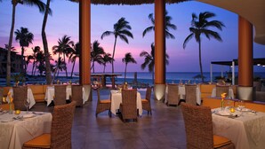 A restaurant with wicker chairs and tables set for dining, overlooking a beach with palm trees and a sunset. - Altitude by Krystal Grand Los Cabos – All Inclusive (San José del Cabo)