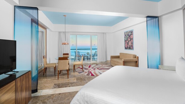 Habitación de hotel con una cama grande, televisión, zona de comedor con vista al océano y balcón con cortinas.