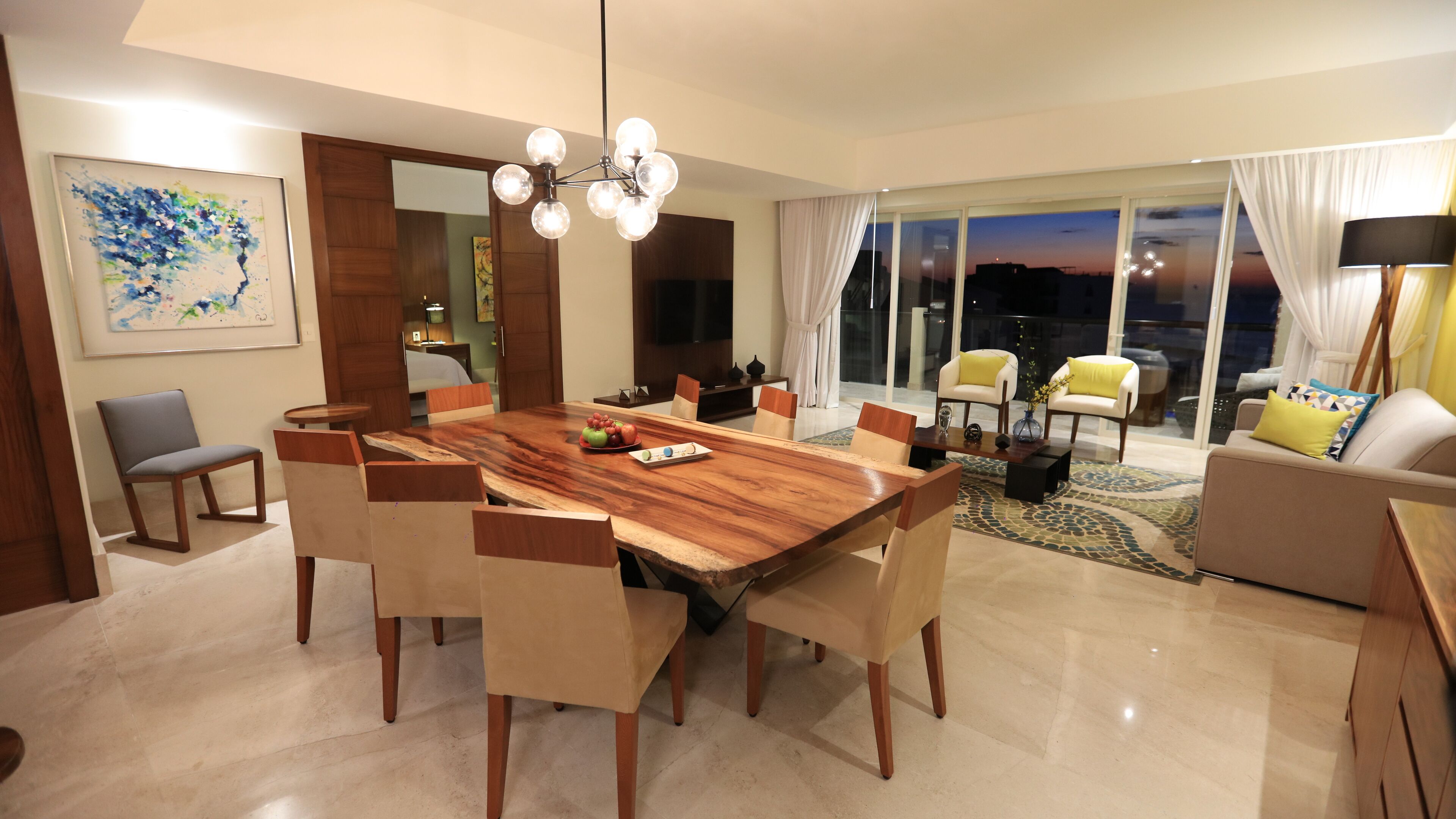 A modern living room with a dining area, featuring a large wooden table, comfortable seating, a TV, and a view of the city at dusk.