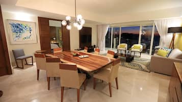 Uma sala de estar moderna com área de jantar, apresentando uma grande mesa de madeira, assentos confortáveis, uma TV e vista da cidade ao entardecer.