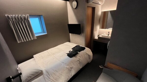 Une chambre d’hôtel de taille réduite, avec un lit simple, une petite fenêtre avec stores, une table de chevet avec une lampe, un miroir et une salle de bain équipée d’un lavabo et d’un miroir.