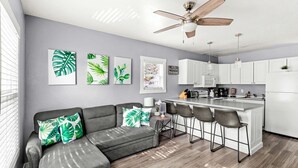 Une cuisine moderne avec un coin bar, un ventilateur de plafond et un canapé gris agrémenté de coussins aux motifs tropicaux.