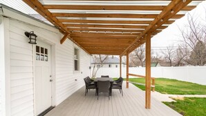 Een overdekt terras met een houten pergola, een tafel met stoelen, en een wit huis op de achtergrond.