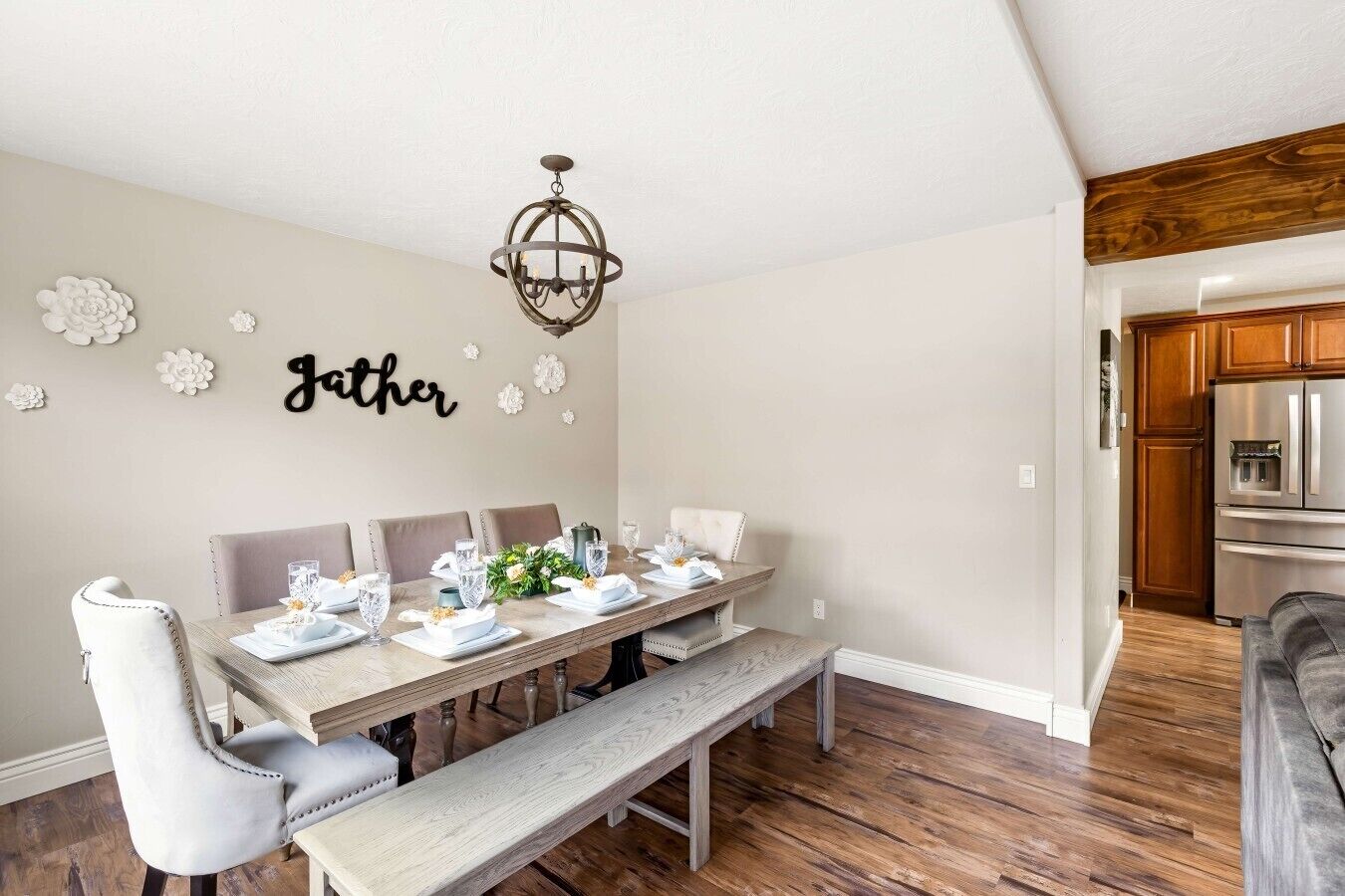 Un espace repas avec une longue table, des chaises blanches, un banc, un lustre et une cuisine en arrière-plan.