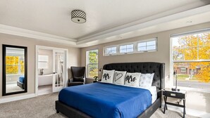 Un dormitorio con una cama grande, una silla, una mesita de noche, una ventana con vistas y una puerta que da a otra habitación.