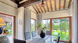 Un comedor con mesa de vidrio, sillas y un florero con flores. Se observa una ventana con cortinas y vistas al verdor exterior.
