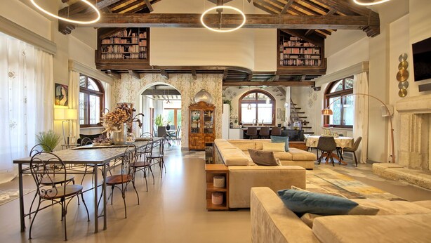 Amplia sala de estar con techo alto, vigas de madera a la vista y una zona de estar central con sofá y sillones. Se observan estanterías llenas de libros y una mesa de comedor con sillas.
