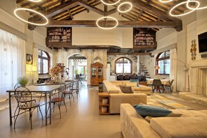 Amplia sala de estar con techo alto, vigas de madera a la vista y una zona de estar central con sofá y sillones. Se observan estanterías llenas de libros y una mesa de comedor con sillas.