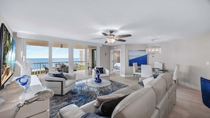 A spacious living room with a large window overlooking the ocean, a ceiling fan, and a dining area with a chandelier. - Beachfront condo with exceptional views – CL0802 (Naples)