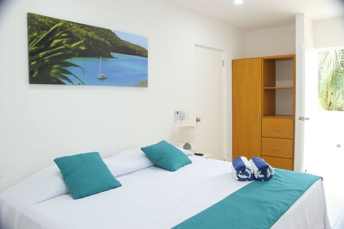 Een netjes opgemaakt bed met teal kussens en een teal sprei, een houten garderobe, een schilderij van een kustlandschap en een klein nachtkastje met een lamp.