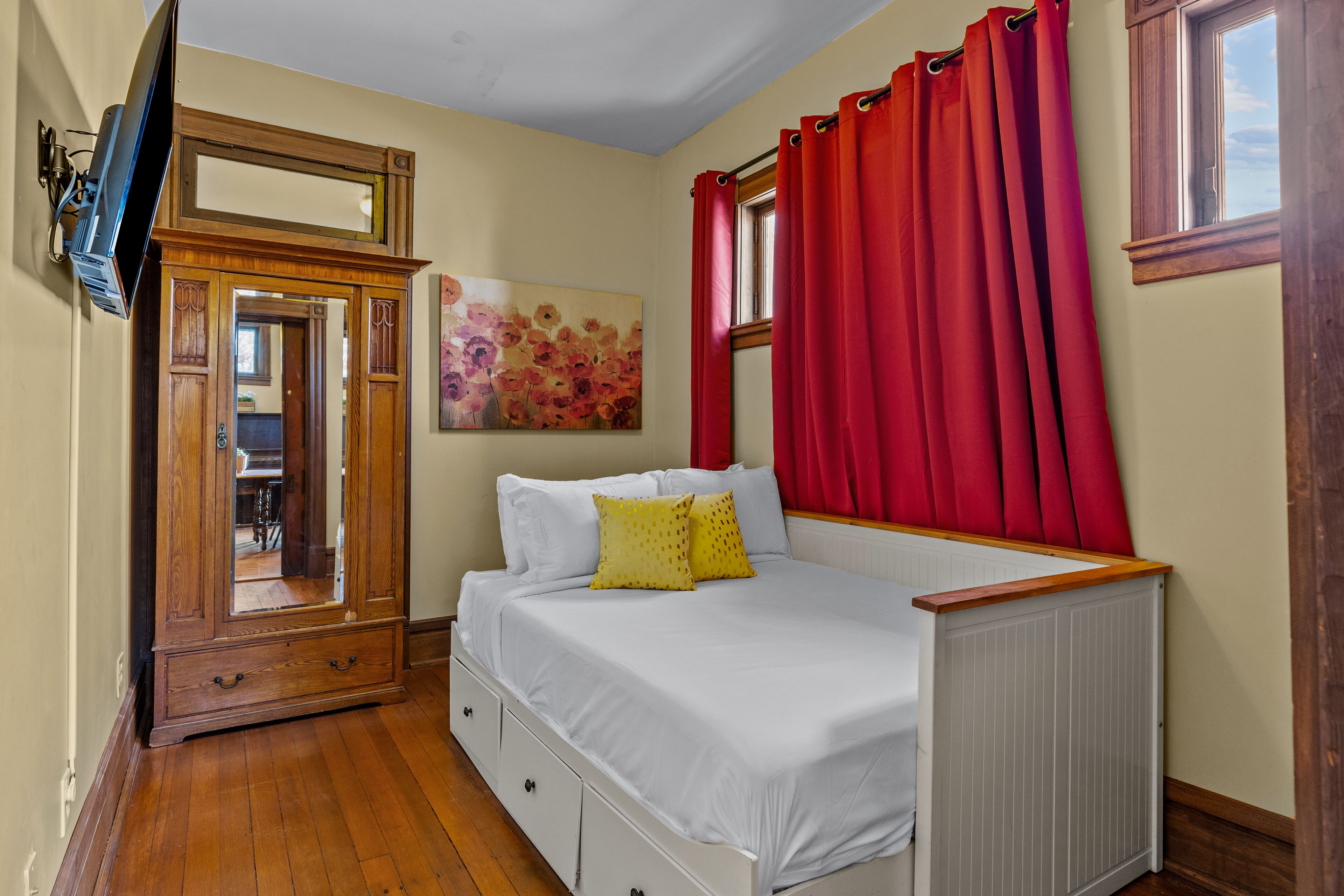 Une chambre à coucher avec un lit, une commode, un miroir, une fenêtre avec des rideaux rouges et un tableau accroché au mur.