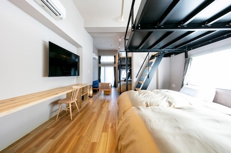 A modern hotel room with a wooden floor, a bed, a desk with a chair, a TV mounted on the wall, and a staircase leading to an upper level.