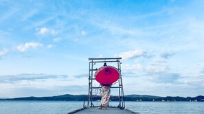 A person standing on a dock with a red umbrella, facing a body of water and distant hills. - Room is up to youOcean view for 3 people vs |  / Nanao Ishikawa (Nanao)