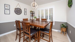 A dining area with a wooden table and chairs, a chandelier, framed pictures, and a window with blinds.