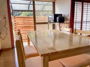 A wooden dining table with cushioned chairs, a television on a stand, and a view of greenery outside.