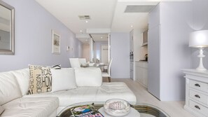 A modern living room with a white sofa, a glass coffee table, and a dining area in the background. - Belle Escapes - Bay View Lookout (Glenelg)