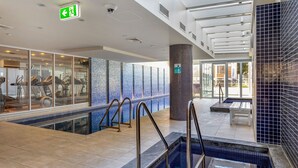 A modern indoor swimming pool area with exercise equipment, a glass wall, and a tiled wall.