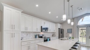 A modern kitchen with white cabinetry, a central island with bar stools, stainless steel appliances, and pendant lighting. - Island Oasis Steps From the Sugar Sand Beach (Fort Myers Beach)