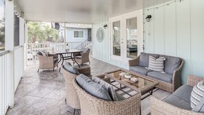 A covered patio area with wicker furniture, a glass coffee table, and a view of a house in the background. - Island Oasis Steps From the Sugar Sand Beach (Fort Myers Beach)