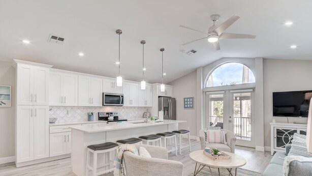 A modern kitchen with white cabinets, a central island with stools, a dining area with a round table, and a living room with a sofa and TV. - Island Oasis Steps From the Sugar Sand Beach (Fort Myers Beach)