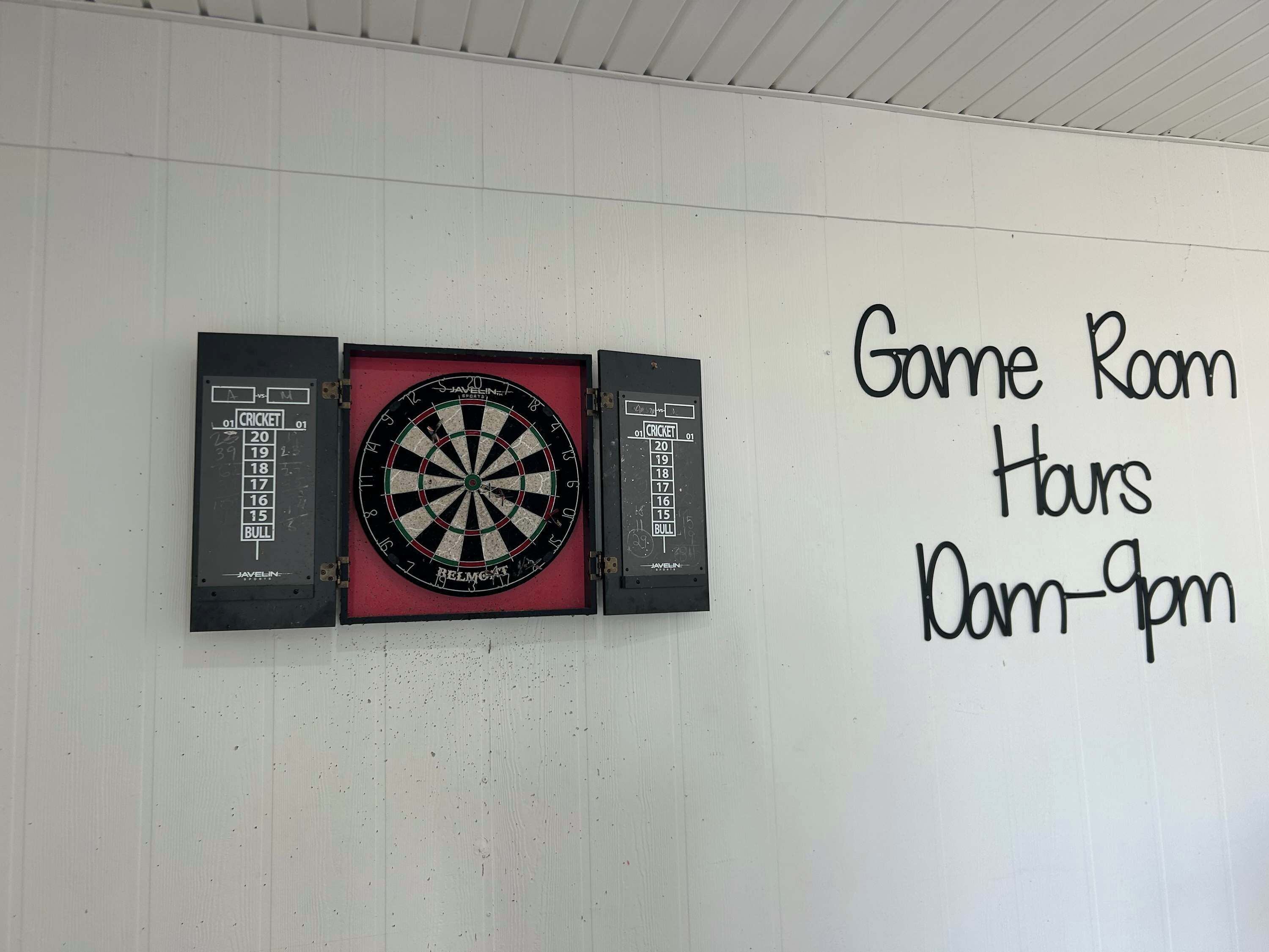 A dartboard mounted on a wall with a sign indicating 'Game Room Hours 10am-9pm'.
