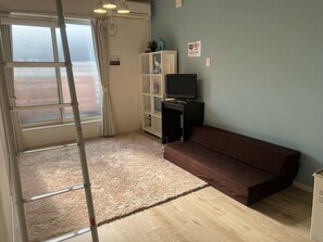 Une pièce avec un canapé marron, un téléviseur posé sur un meuble, une étagère blanche, un plancher en bois et une grande fenêtre avec des rideaux.