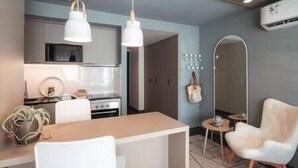 Een moderne keuken met witte kasten, een fornuis en hanglampen. Er is een eethoek met een houten tafel en witte stoelen. Ook zichtbaar is een spiegel en een klein rond tafeltje met een lamp.