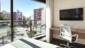 Hotelkamer met een groot raam met uitzicht op de stad, een bed, een bureau met stoel en een wandgemonteerde flatscreen-tv.
