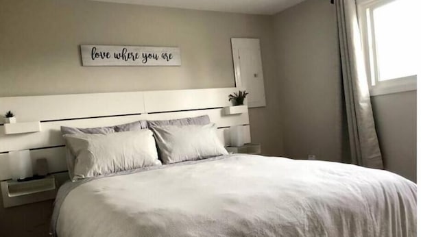 A bedroom with a bed, a bedside table, a nightstand, a wall-mounted shelf, and a window with curtains. - Comfy and modern private suite - everything you need for a short to midterm stay (Dryden)