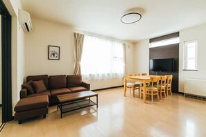 A living room with a brown sofa, a coffee table, and a dining table with chairs. There is a TV mounted on the wall and an air conditioning unit on the wall.